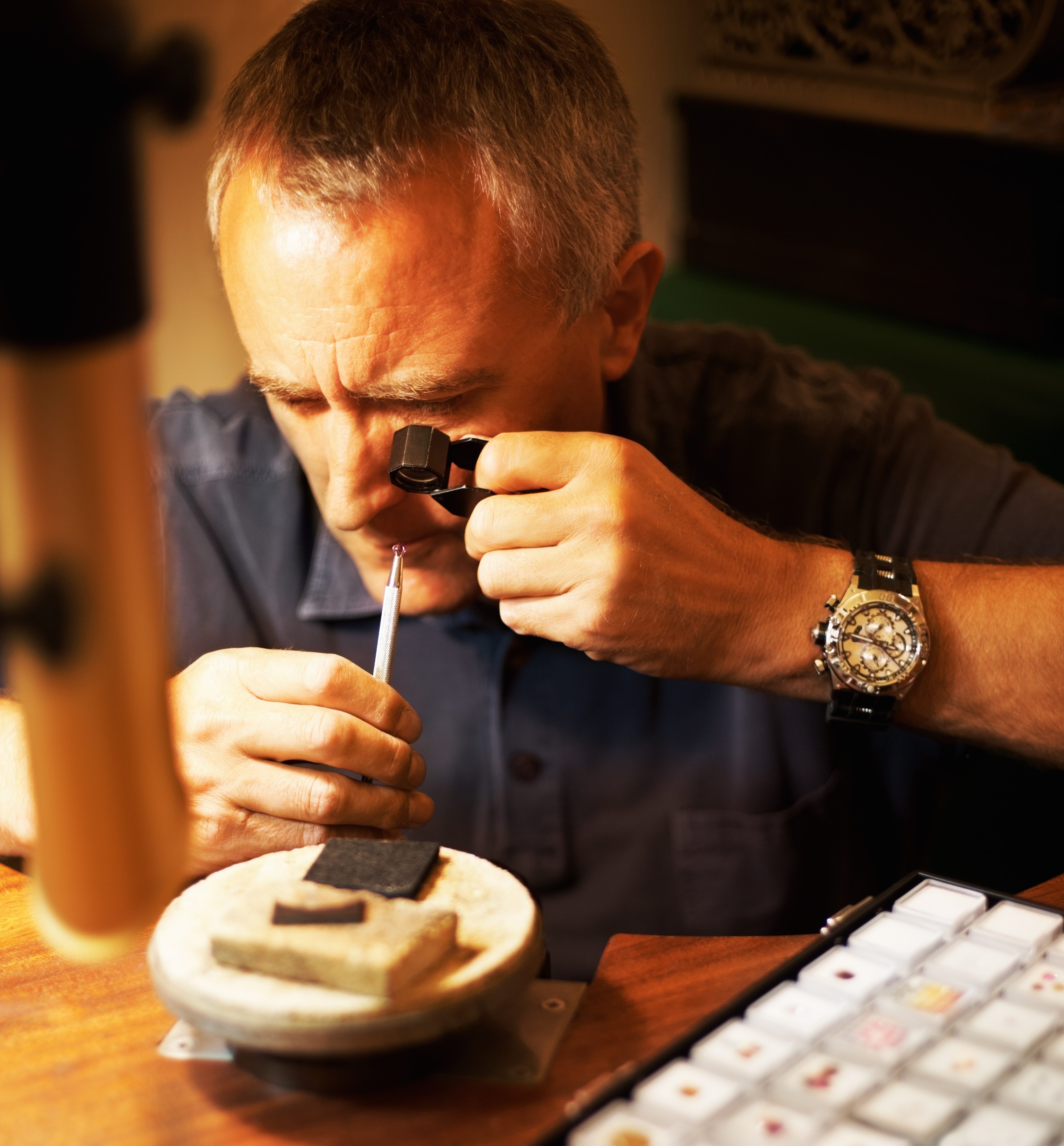 Man inspecting a gemstone using a jeweler’s loupe at a workbench. He is holding the stone with precision tweezers, focused intently, with various gem tools and samples laid out in front of him. The setting appears to be a gemologist’s or jeweler’s workspace, lit warmly.