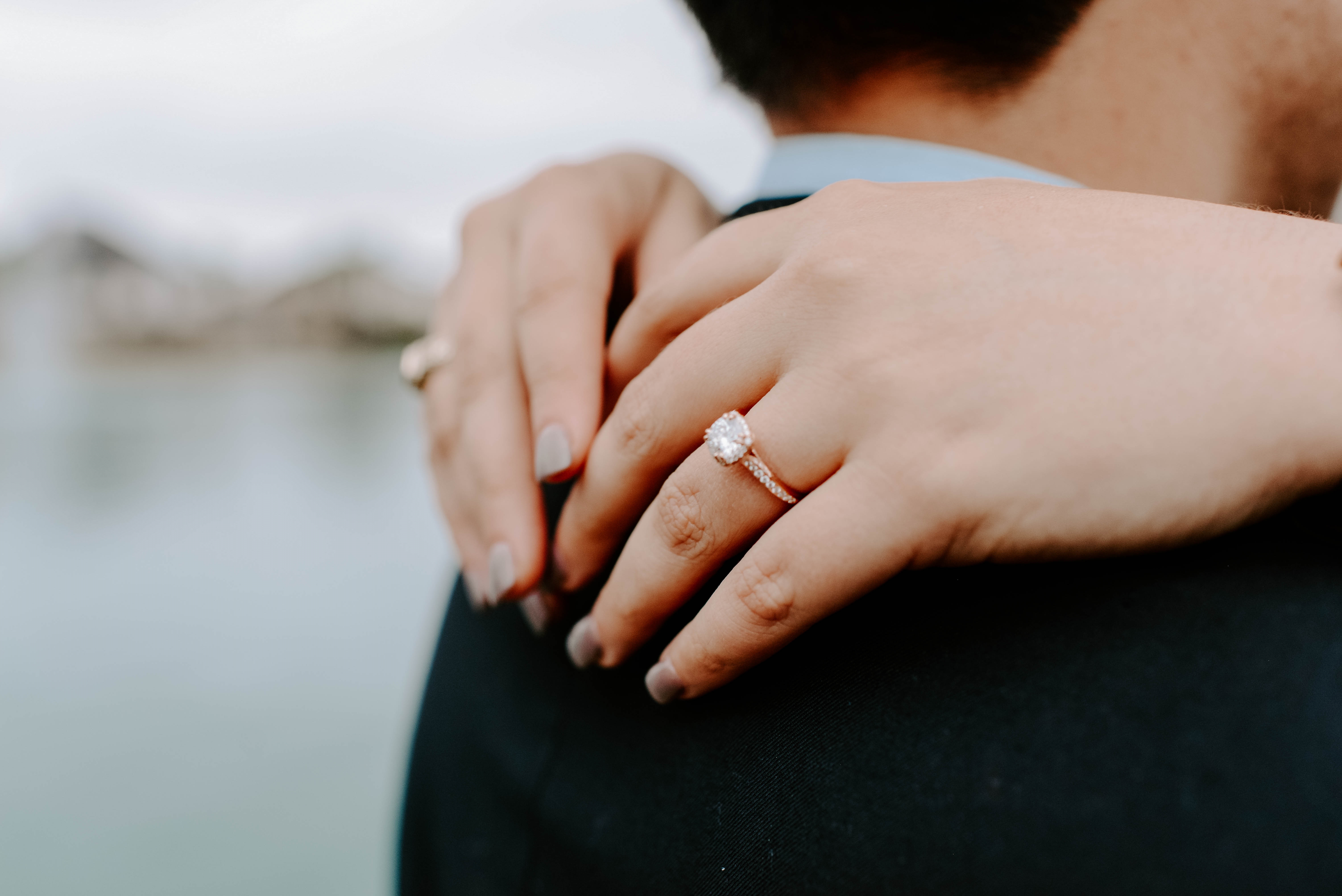 Close-up of a woman’s hands resting on her partner’s shoulder, showcasing a diamond engagement ring with a pavé band